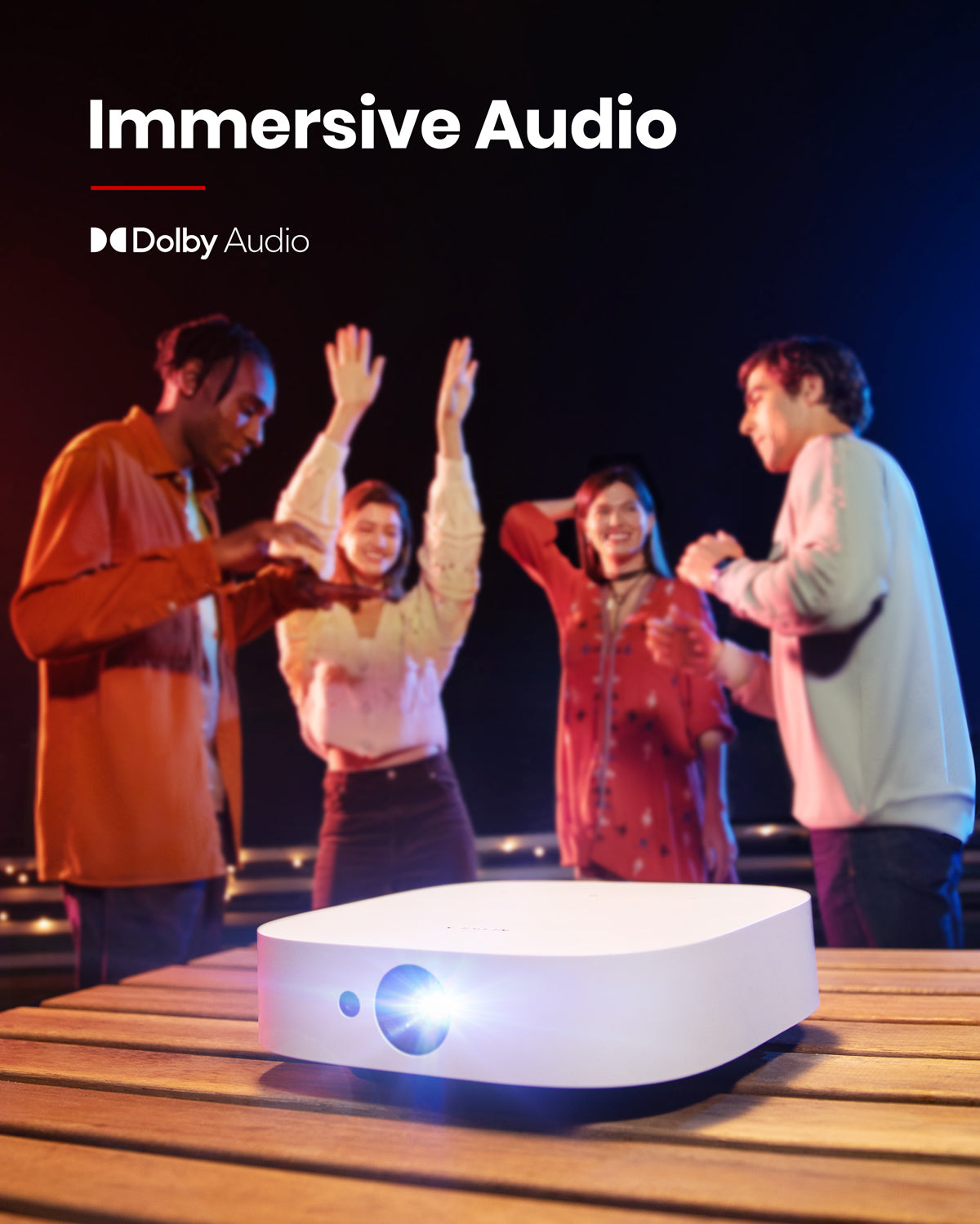 A group of friends dance at night while a Nebula Solar Portable projector on top of a wooden table emits a blue light.